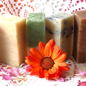 May include: Six bars of handmade soap in various colors, including purple, cream, green, white, brown, and yellow, arranged in a row on a white lace tablecloth with an orange flower in front.