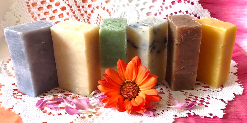 May include: Six bars of handmade soap in various colors, including purple, cream, green, white, brown, and yellow, arranged in a row on a white lace tablecloth with an orange flower in front.