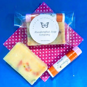 May include: A set of three handcrafted soap products from the Handcrafted Soap Company. The set includes two bars of soap and a lip balm. The soap bars are yellow and orange with a swirl design. The lip balm is pink and orange with a butterfly design. The products are packaged in clear plastic bags.