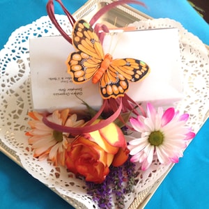 May include: A white gift box with a handwritten message, decorated with a butterfly, a rose, and daisies on a white doily. The message reads "Olive Oil Organics,  Sampler Box,  Made with Love."
