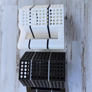 Puede incluir: Se muestran dos contenedores de almacenamiento geométricos, uno blanco y otro negro. Cada uno tiene una forma rectangular con un patrón de panal perforado. Bandas negras envuelven los contenedores. Los contenedores están sobre una superficie de vetas de madera clara.