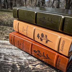 May include: A stack of three leather-bound books with embossed titles and floral designs. The books are in shades of green, tan, and brown, with the titles "Emma", "Sense & Sensibility", and "Pride & Prejudice" visible. The author's name "Austen" is also embossed.