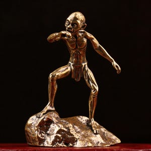 May include: A bronze-colored figurine of a creature, standing on a rock. The figure is in a dynamic pose, with one arm raised and the other extended. The creature has a gaunt appearance, with detailed musculature and a loincloth. The background is dark.