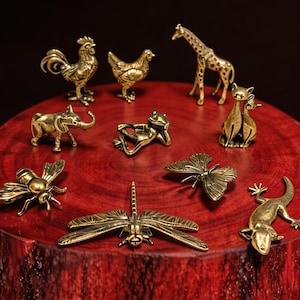 May include: A collection of small, detailed brass animal figurines, including a rooster, hen, giraffe, elephant, frog, cats, bee, butterfly, dragonfly, and lizard. The figurines are displayed on a red wooden surface against a dark background.