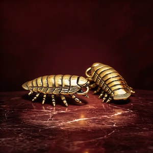 Puede incluir: Dos esculturas metálicas doradas detalladas de cochinillas, con cuerpos segmentados y múltiples patas. Se exhiben sobre una superficie de mármol rojo, contra un fondo rojo oscuro.