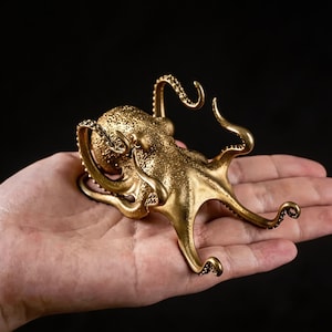Op de afbeelding: Een gedetailleerde, gouden octopussculptuur met een getextureerde huid en gekrulde tentakels. De octopus is een decoratief item, waarschijnlijk gemaakt van metaal, en wordt tegen een donkere achtergrond getoond.