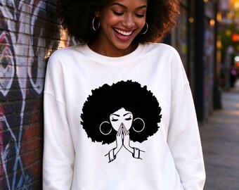 May include: A white long-sleeve sweatshirt featuring a black graphic print of a person with a large afro hairstyle and hoop earrings, with hands pressed together in a prayer position centered on the front of the garment.