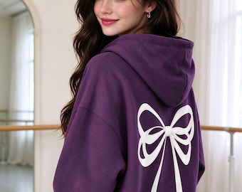 May include: A deep purple hooded sweatshirt featuring a large, white graphic print of a bow on the back. The garment has long sleeves, a hood, and a relaxed fit, showcasing the back design clearly while worn in a casual setting.