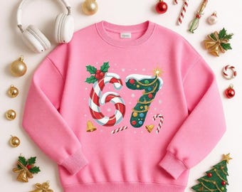May include: A pink long-sleeve sweatshirt featuring a graphic of the number 67, where the 6 is a red and white candy cane and the 7 is a decorated green Christmas tree. The design includes holly leaves, berries, and small candy canes.