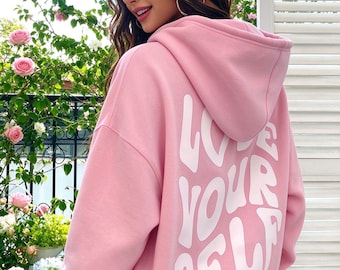 May include: A light pink hooded sweatshirt featuring the text LOVE YOUR SELF printed in large, white, wavy-style lettering on the back. The garment includes a hood, long sleeves, and ribbed cuffs, showcasing a casual design with a bold graphic.