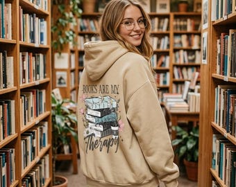 May include: A beige hooded sweatshirt featuring a graphic on the back with a stack of books, flowers, and the text "BOOKS ARE MY Therapy". The hoodie is worn with light blue denim jeans. The design includes illustrations of books, floral accents, and stars.