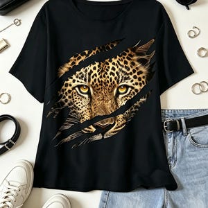 May include: A black short-sleeved t-shirt featuring a graphic print of a leopard face appearing through three diagonal claw marks. The leopard has detailed fur patterns and piercing yellow eyes. The shirt is displayed with accessories and jeans.
