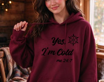 May include: A maroon hooded sweatshirt featuring a front kangaroo pocket and matching drawstrings. The chest displays black text that reads "Yes, I'm Cold" above the text "me 24:7" and a black snowflake graphic icon.