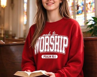 May include: A red long-sleeve crewneck sweatshirt featuring the white text "made to WORSHIP" and "PSALM 95:1" printed on the front. The garment is paired with blue denim jeans and is worn while holding an open book.