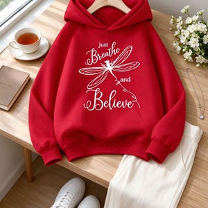 May include: A bright red hooded sweatshirt featuring a white graphic of a dragonfly and the text "just Breathe and Believe" printed on the front. The hoodie has long sleeves, a front kangaroo pocket, and ribbed cuffs and hem for a structured fit.