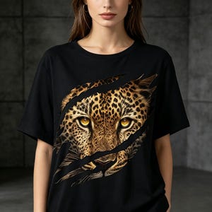 May include: A black short-sleeved t-shirt featuring a graphic print of a leopard face appearing through a series of diagonal claw marks. The leopard has detailed golden-brown fur with black spots and piercing yellow eyes centered on the chest.