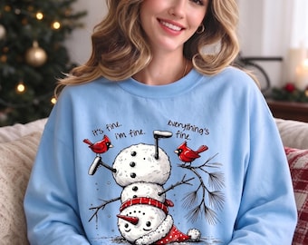 May include: A light blue long-sleeve sweatshirt featuring a graphic of an upside-down snowman wearing a red hat and scarf, two red cardinals, and the text "it's fine. i'm fine. everything's fine." printed in black above the snowman illustration.