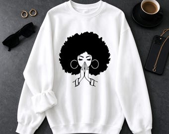 May include: A white long-sleeve crewneck sweatshirt featuring a black graphic print of a person with a large afro hairstyle and hoop earrings, shown with eyes closed and hands pressed together in a prayer position at the center chest.