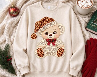 May include: A beige crewneck sweatshirt featuring a central graphic of a teddy bear wearing a leopard print hat and lace accents. The bear has a red bow with holly berries at its neck. The leopard print pattern is also on the bear's feet.