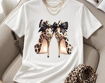 May include: A white short-sleeved t-shirt featuring a graphic print of two leopard print high-heeled shoes. Each shoe is adorned with a large black bow at the heel and intricate gold-toned embellishments near the stiletto base.