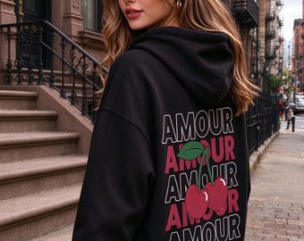 May include: A black hooded sweatshirt featuring a graphic on the back with the word AMOUR repeated five times in alternating white and dark red text, centered with a pair of red cherries with a green leaf positioned over the middle word.