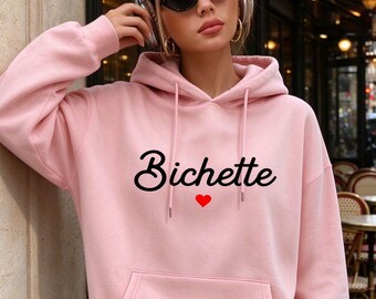 May include: A light pink hooded sweatshirt featuring long sleeves, a front kangaroo pocket, and matching drawstrings. The center of the chest displays the word "Bichette" in black cursive script lettering above a small, solid red heart icon.