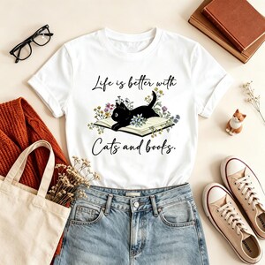 Puede incluir: Una camiseta blanca de manga corta con un gráfico de un gato negro tumbado sobre un libro abierto rodeado de flores silvestres de colores. El texto "Life is better with Cats and books." está impreso en negro sobre y bajo la ilustración.