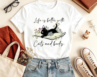 May include: A white short-sleeved t-shirt featuring a graphic of a black cat lying on an open book surrounded by colorful wildflowers. The text "Life is better with Cats and books." is printed in black script above and below the illustration.