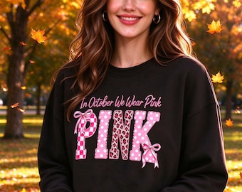 May include: A black long-sleeve sweatshirt featuring the text In October We Wear Pink in white and pink lettering. The word PINK is stylized with a pink and white checkered pattern, leopard print, and polka dots, accented by pink ribbons.