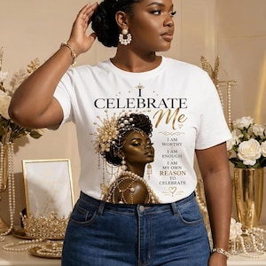 Può includere: Una t-shirt bianca a maniche corte con la grafica di una persona dalla pelle scura che indossa un copricapo e gioielli dorati decorati. Il testo recita: I CELEBRATE Me, I AM WORTHY, I AM ENOUGH, I AM MY OWN REASON TO CELEBRATE. Abbinata a un paio di jeans blu.