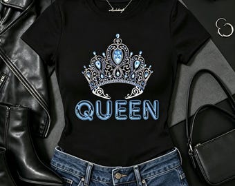 May include: A black short-sleeved t-shirt featuring a graphic of a silver crown adorned with light blue gemstones. Below the crown, the word QUEEN is printed in a bold, light blue font with a dark blue outline, centered on the chest.