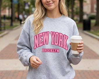 May include: A heather gray crewneck sweatshirt featuring the text NEW YORK in a large, arched pink font with a dark pink outline, and the word BROOKLYN in a smaller, straight pink font underneath. The sweatshirt has long sleeves and a ribbed collar.