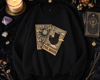 May include: A black crewneck sweatshirt featuring a graphic print of two tarot cards. One card displays a sun and clouds, while the other card shows a black cat and the text THE MOON. The sweatshirt has long sleeves and a ribbed collar.