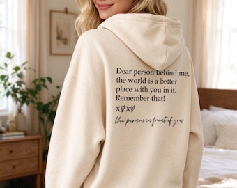 May include: A cream-colored hooded sweatshirt featuring black text on the back that reads: Dear person behind me, the world is a better place with you in it. Remember that! XOXO the person in front of you. The hoodie has long sleeves and a hood.