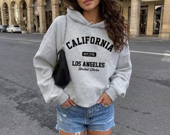 May include: A light gray hooded sweatshirt featuring black text that reads CALIFORNIA, EST. 1776, LOS ANGELES, and United States. The garment has long sleeves, a front kangaroo pocket, and is paired with blue denim shorts and a black shoulder bag.