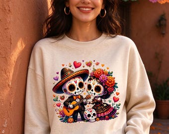 May include: A beige crewneck sweatshirt featuring a colorful graphic print of two cartoon sugar skull characters. One character wears a sombrero and plays a guitar, while the other wears a floral crown. Small hearts and flowers surround them.