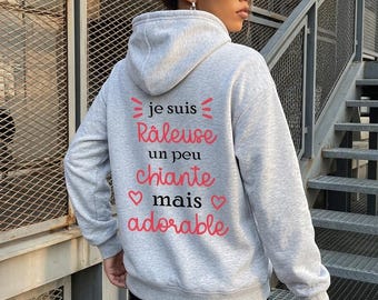 May include: A light gray hooded sweatshirt featuring black and pink text on the back that reads: je suis Râleuse un peu chiante mais adorable, accented with small pink heart symbols. The hoodie has long sleeves and a hood.