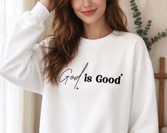 May include: A white long-sleeve crewneck sweatshirt featuring the black text God is Good printed on the front. The word God is written in a cursive script font, while the words is Good are in a bold sans-serif font with a small star icon.
