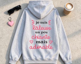 May include: A light gray hooded sweatshirt featuring the French text "je suis Râleuse un peu chiante mais adorable" in black and pink lettering, accompanied by pink heart icons, black sunglasses, and a pink heart keychain.