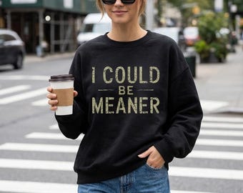 May include: A black long-sleeve crewneck sweatshirt featuring the text I COULD BE MEANER in a distressed, light-colored font across the chest. The garment has a relaxed fit with ribbed cuffs and a ribbed neckline, worn with blue denim jeans.