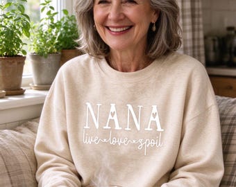May include: A beige crewneck sweatshirt featuring the white text NANA in a bold font with a small heart cutout in the letter A, followed by the cursive white text live love spoil with small heart accents between each word.