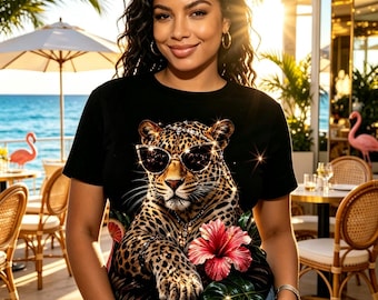 May include: A black short-sleeved t-shirt featuring a graphic print of a leopard wearing cat-eye sunglasses and a jeweled headband. The leopard is surrounded by a large pink hibiscus flower, green leaves, and a slice of citrus fruit.