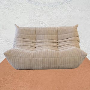 Puede incluir: Sillón de pana beige con un perfil bajo y forma redondeada. La silla tiene una superficie texturizada y acanalada, con un aspecto suave y acogedor. El diseño presenta un asiento ancho y un respaldo con dos pequeñas secciones verticales.