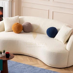 Puede incluir: Un sofá curvo de color crema con una tela texturizada. Cuenta con dos cojines rectangulares y tres cojines redondos en crema, naranja, morado y azul marino. Una pequeña mesa auxiliar redonda y una alfombra azul están en primer plano.