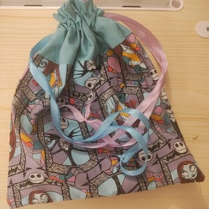 May include: A drawstring bag featuring a patterned fabric with characters from a popular animated film. The bag has a light blue top and is tied with blue and pink ribbons. The main fabric has a purple background.