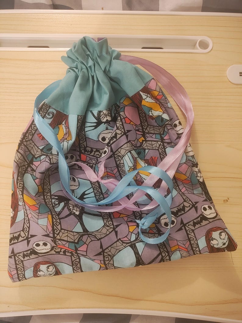 May include: A drawstring bag featuring a patterned fabric with characters from a popular animated film. The bag has a light blue top and is tied with blue and pink ribbons. The main fabric has a purple background.