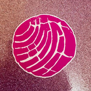 May include: A bright pink sticker in the shape of a concha, a traditional Mexican sweet bread. The sticker has a white pattern on the pink background, and is set against a glittery pink background.