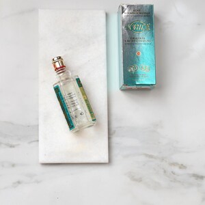 May include: A clear glass bottle of 4711 Original Eau de Cologne with a red and gold cap, resting on a white marble slab. A teal and silver box of the cologne is next to the bottle. The box has the text "4711" and "Original Eau de Cologne".