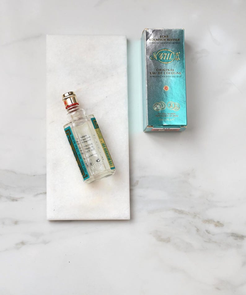 May include: A clear glass bottle of 4711 Original Eau de Cologne with a red and gold cap, resting on a white marble slab. A teal and silver box of the cologne is next to the bottle. The box has the text "4711" and "Original Eau de Cologne".