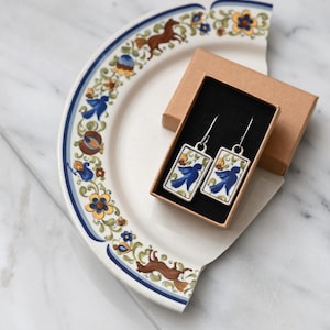 Op de afbeelding: Zilverkleurige oorbellen met blauw-witte bloempatronen in een klein bruin doosje. De oorbellen liggen op een gebroken witte plaat met een blauw-witte bloemenrand. De plaat heeft bruine en blauwe bloemen- en diermotieven.